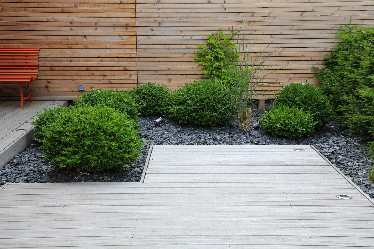 Aménagement paysager extérieur moderne avec terrasse en bois composite, jardin minéral composé de galets anthracite, arbustes verts taillés en boules, graminées ornementales et bambous