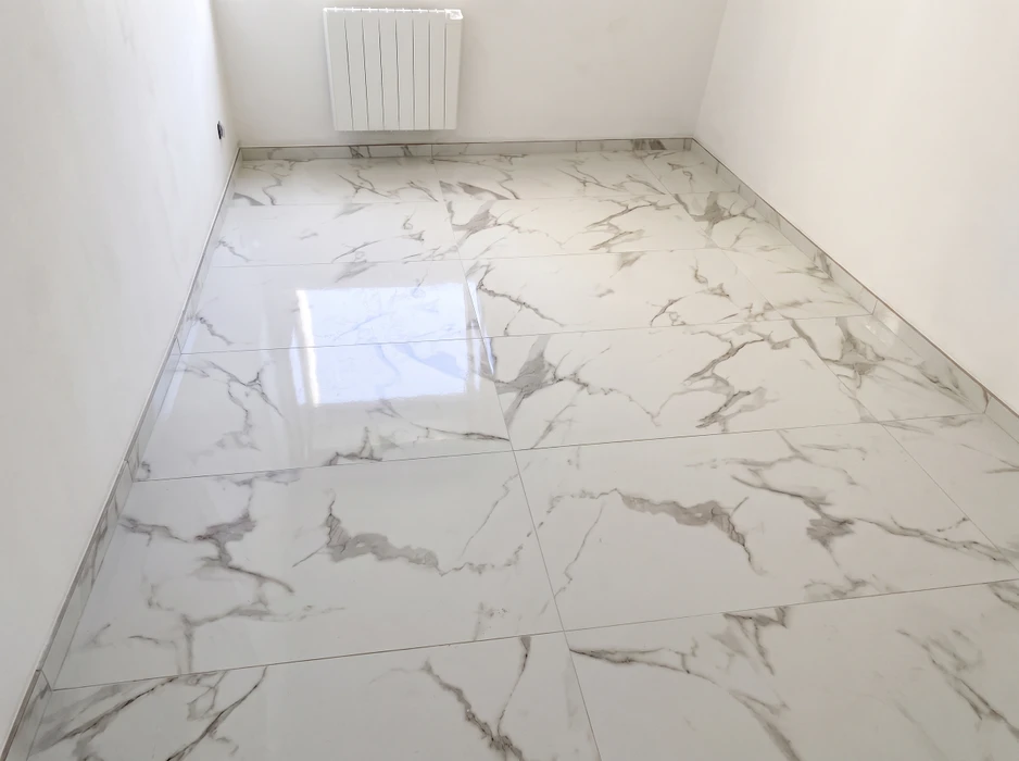 Travailleur en train de poser du carrelage sur sol intérieur, à genoux, portant des gants de protection jaunes et bleus